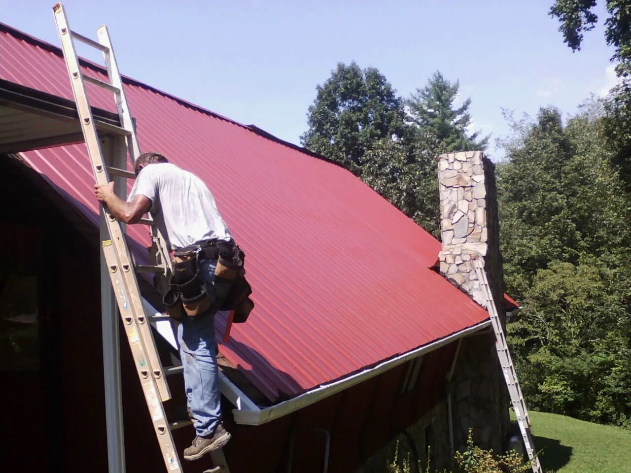 Roofer on a ladder beside a red metal roof and stone chimney — Soffit & Fascia Repair by Roofing Brothers in Somonauk